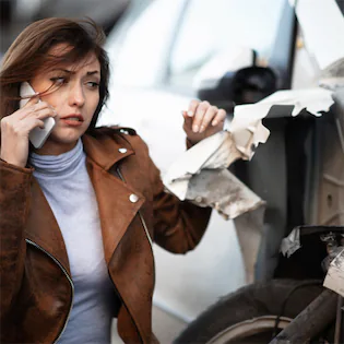 Woman sitting beside a broken car, making a call, reflecting concerns about the legality of driving a crashed car in California